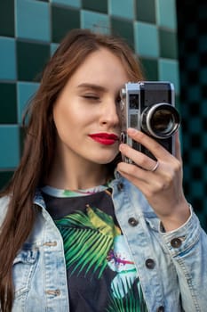 Cool girl model with retro film camera wearing a denim jacket, dark hair outdoors over city wall in a cage background. Having fun in the city with camera, travel photo of photographer.