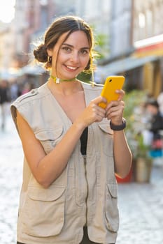 Smiling young woman using smartphone typing text messages browsing internet, finishing work, looking at camera outdoors. Girl tourist walking in urban sunshine city street. Town lifestyles. Vertical