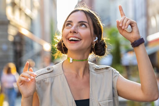 Happy relaxed overjoyed pretty young woman wearing wireless earphones choosing, listening favorite energetic disco music dancing outdoors. Girl walking alone in urban city sunshine street background
