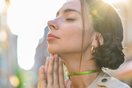 Portrait of pretty young woman praying with closed eyes to God asking for blessing help, forgiveness, church outdoors. Pretty girl clasping hands closed eyes wishing luck in city street. Town religion