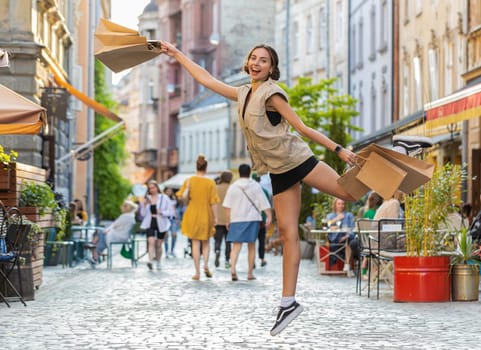Joyful smiling pretty young woman shopaholic consumer after shopping sale with full bags with gifts jumping outdoors. Girl tourist traveler walking along the urban city street road. Town lifestyles