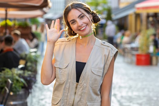 Portrait of young woman smiling friendly at camera, waving hands gesturing hello, hi, greeting or goodbye, welcoming with hospitable expression outdoors. Girl tourist walking on urban city town street