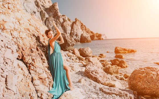 Side view a Young beautiful sensual woman in a red long dress posing on a rock high above the sea during sunrise. Girl on the nature on blue sky background. Fashion photo.