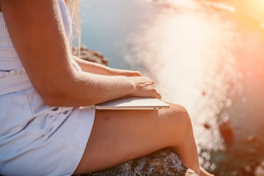 Successful business woman in yellow hat working on laptop by the sea. Pretty lady typing on computer at summer day outdoors. Freelance, travel and holidays concept.