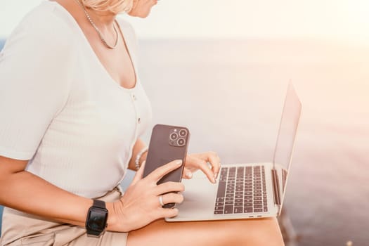 Digital nomad, Business woman working on laptop by the sea. Pretty lady typing on computer by the sea at sunset, makes a business transaction online from a distance. Freelance remote work on vacation