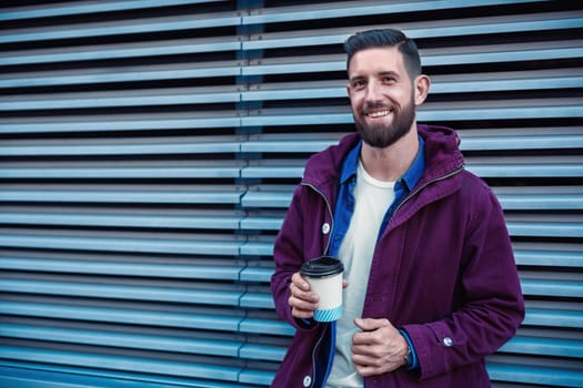 Outdoor fall or winter portrait of handsome hipster man with beard, white t-shirt, blue shirt and maroon jacket holding cup of hot coffee. Ribbed urban wall background.