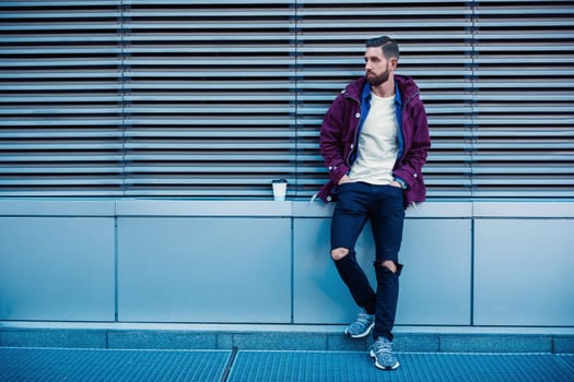 Outdoor fall or winter portrait of handsome hipster man with beard, white t-shirt, blue shirt and maroon jacket holding cup of hot coffee. Ribbed urban wall background.