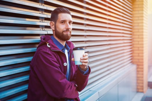 Outdoor fall or winter portrait of handsome hipster man with beard, white t-shirt, blue shirt and maroon jacket holding cup of hot coffee. Ribbed urban wall background.