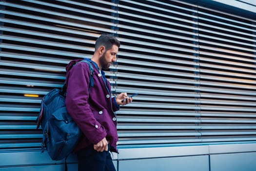 Confident bearded man holding in and mobile phone while standing near street wall in sunny summer evening. Young fashionable hipster guy using cell telephone while posing outdoors near copy space area