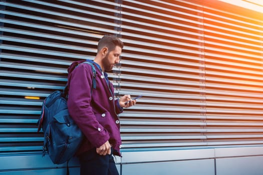 Outdoor fall or winter portrait of handsome hipster man with beard, white t-shirt, blue shirt and maroon jacket holding smartphone. Ribbed urban wall background.