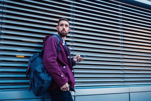 Confident bearded man holding in and mobile phone while standing near street wall in sunny summer evening. Young fashionable hipster guy using cell telephone while posing outdoors near copy space area