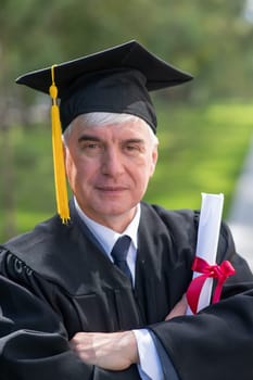 Portrait of an elderly man in a graduation gown and with a diploma in his hands outdoors. Vertical