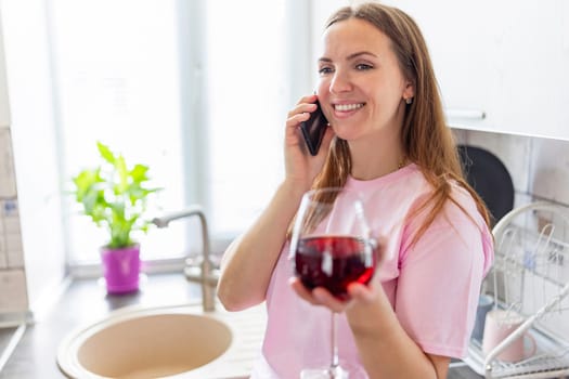 Happy relaxed young woman standing in kitchen with glass of red wine and using her smartphone.