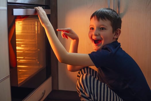 A child is sitting near the oven in the kitchen and waiting. Curious boy is watching through the glass of kitchen oven. Baking pizza, muffins , cupcakes or cookies.