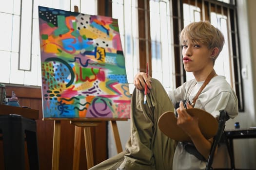 Playful asian gay male artist sitting front of easel in bright art studio and looking at camera.