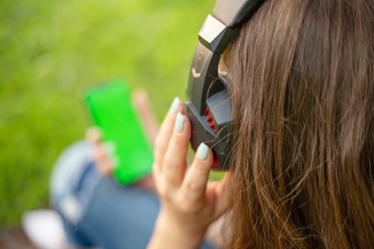 A woman is holding a smartphone and listening to music on headphones while sitting on the grass in a park.