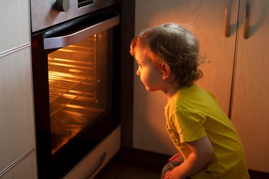 A child is sitting near the oven in the kitchen and waiting. Curious boy is watching through the glass of kitchen oven. Baking pizza, muffins , cupcakes or cookies.