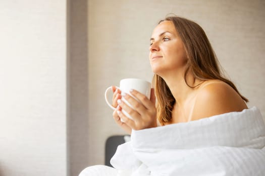 Beautiful young woman drinking a cup of coffee or tea while sitting on the bed after waking up in the morning. A woman is enjoying her drink in bed at home.