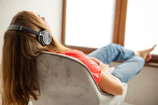 Young beautiful woman sitting in armchair near a window and listening to music using headphones. Happy calm woman in earphones rest on cozy armchair at home , stress free concept.