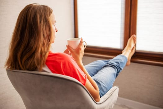 Young beautiful woman in casual wear sitting in armchair with cup of hot coffee. Happy woman relaxing in armchair near a window.