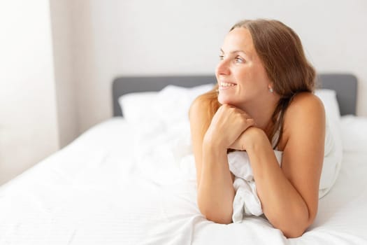 Young smiling attractive brunette woman lies in a big white bed.