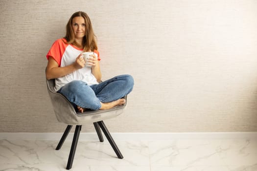 Young beautiful woman in casual wear sitting in armchair with cup of hot coffee. Happy woman relaxing in armchair.