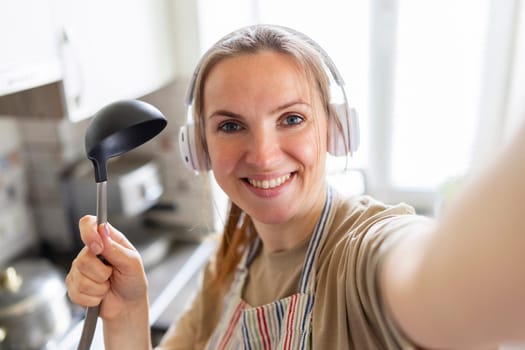 Cheerful young housewife in apron and earphones with soup ladle taking selfie photo in cozy kitchen.