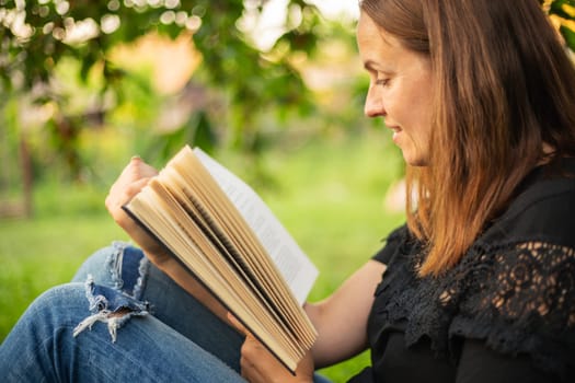 A woman sits near a tree in the park and holds a book in her hands. A woman in jeans and a t-shirt reading a book outdoor.