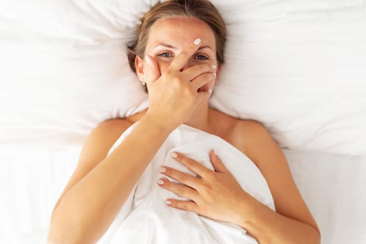 Top view of happy young woman peeking through her fingers and smiling while lying in bed.