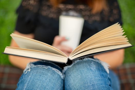 A woman sits near a tree in the park and holds a book and a cup with a hot drink in her hands. A woman in jeans and a t-shirt reading a book outdoor.