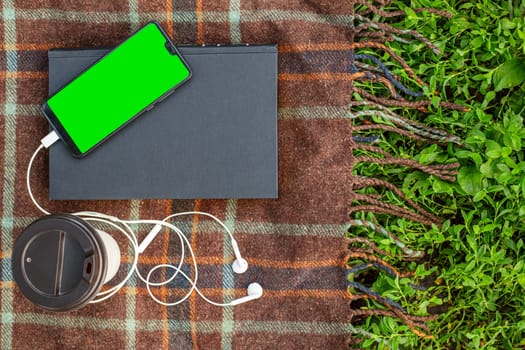 Cup of coffee, book and smartphone with headphones on blanket in the park or garden.