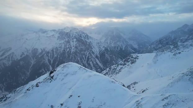 Mystical dawn with clouds in snowy mountains. The white hills are covered with clouds and snow. Steep rocky cliffs. The high peaks of the peaks. The rays of the sun at dawn pass through the clouds