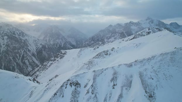 Mystical dawn with clouds in snowy mountains. The white hills are covered with clouds and snow. Steep rocky cliffs. The high peaks of the peaks. The rays of the sun at dawn pass through the clouds