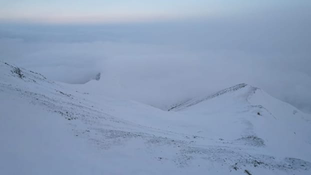 Mystical dawn with clouds in snowy mountains. The white hills are covered with clouds and snow. Steep rocky cliffs. The high peaks of the peaks. The rays of the sun at dawn pass through the clouds