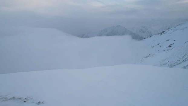 Mystical dawn with clouds in snowy mountains. The white hills are covered with clouds and snow. Steep rocky cliffs. The high peaks of the peaks. The rays of the sun at dawn pass through the clouds