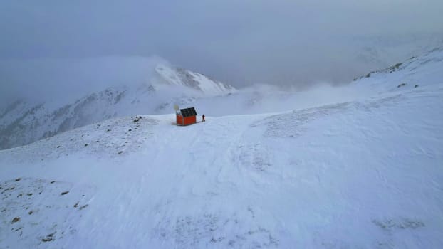 A rescue hut high in the mountains among the clouds. There is a climber standing next to the house. White clouds enveloped the snowy mountains. Yellow dawn and clouds. Snowy hills. 01.05.2023 Almaty