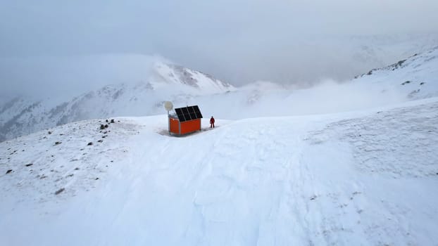 A rescue hut high in the mountains among the clouds. There is a climber standing next to the house. White clouds enveloped the snowy mountains. Yellow dawn and clouds. Snowy hills. 01.05.2023 Almaty