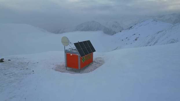 A rescue hut high in the mountains among the clouds. There is a climber standing next to the house. White clouds enveloped the snowy mountains. Yellow dawn and clouds. Snowy hills, stones lie. Almaty