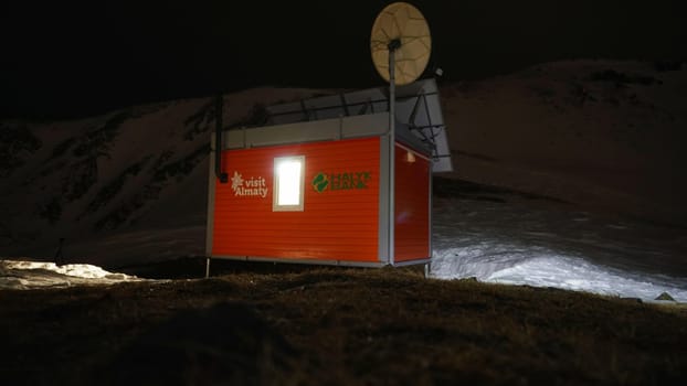 Modern rescue hut in the mountains at night. The van is equipped with rescue equipment. Installed satellite dish, oven, chimney, light, insulated. Winter, lots of snow. A house high in the mountains