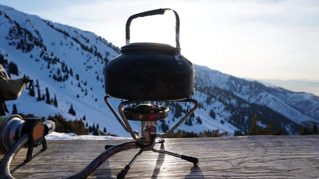 The burner is burning with a blue fire with a kettle. The burner and the cylinder are on the bench. In the background there are green fir trees, mountains in white snow and a beautiful sunset