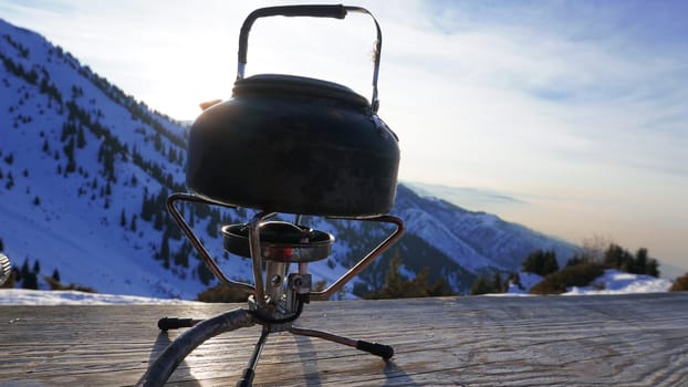 The burner is burning with a blue fire with a kettle. The burner and the cylinder are on the bench. In the background there are green fir trees, mountains in white snow and a beautiful sunset