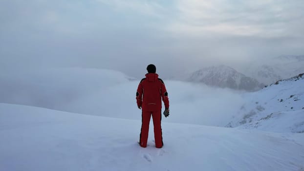 A climber in a red suit stands at the top of the mountain. Looks at the heavenly ocean of white clouds. There is snow all around, blue sky, dawn and calm. An epic moment of life. There are tours