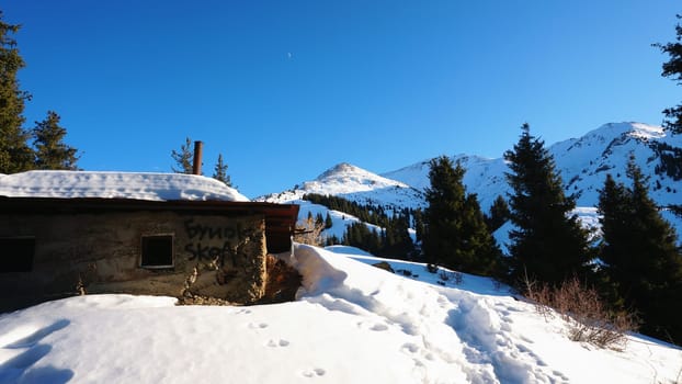 An abandoned house in the snowy mountains. Christmas trees and bushes grow. Clear sky and moon. A small moon in the distance above the top of the peak. There is a lot of snow on the roof. Almaty