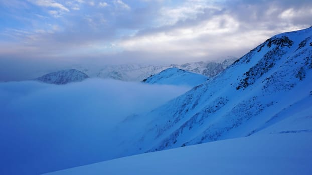 An ocean of clouds in the snowy mountains at dawn. White clouds are like a carpet in a gorge. Waves rise and descend from the mountains. Sunrise of the yellow sun. Clear sky, mountain peaks. Almaty