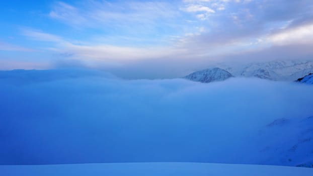 An ocean of clouds in the snowy mountains at dawn. White clouds are like a carpet in a gorge. Waves rise and descend from the mountains. Sunrise of the yellow sun. Clear sky, mountain peaks. Almaty