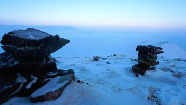 An ocean of clouds in the snowy mountains at dawn. White clouds are like a carpet in a gorge. Waves rise and descend from the mountains. Sunrise of the yellow sun. There are signposting stones. Almaty