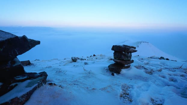 An ocean of clouds in the snowy mountains at dawn. White clouds are like a carpet in a gorge. Waves rise and descend from the mountains. Sunrise of the yellow sun. There are signposting stones. Almaty