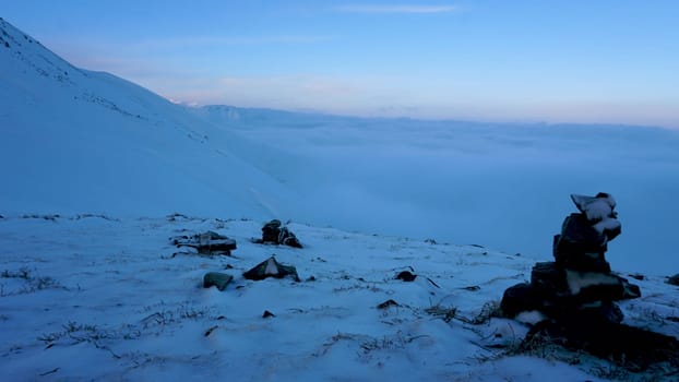 An ocean of clouds in the snowy mountains at dawn. White clouds are like a carpet in a gorge. Waves rise and descend from the mountains. Sunrise of the yellow sun. There are signposting stones. Almaty