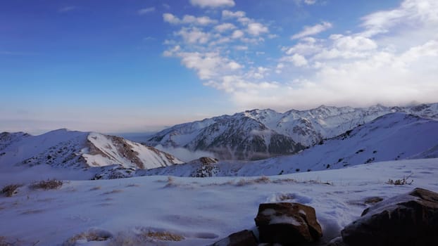 An ocean of clouds in the snowy mountains at dawn. White clouds are like a carpet in a gorge. Waves rise and descend from the mountains. Sunrise of the yellow sun. There are stones lying. Kazakhstan