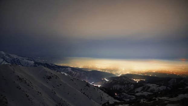 View of the night city from the snowy mountains. The glow from the city rises to the clouds. Bright lights are visible. In places, even the stars are visible. Steep cliffs, big rocks and snow. Almaty